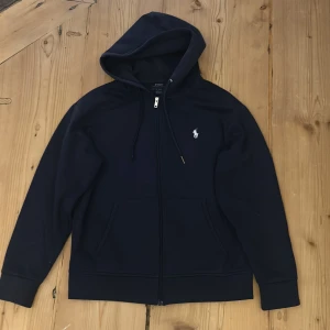 Marinblå hoodie från Polo Ralph Lauren - Snygg marinblå hoodie från Polo Ralph Lauren med dragkedja och huva. Tröjan har snörning i huvan, två fickor fram och den klassiska vita Polo-loggan broderad på bröstet. Perfekt för chill dagar och enkel att matcha med jeans eller joggers.