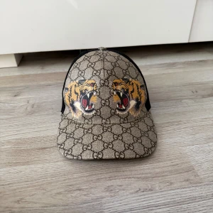 Gucci keps med tigertryck och GG-mönster - Snygg keps från Gucci med klassiskt GG-monogram i beige och brunt, prydd med två broderade tigrar framtill. Baksidan har svart mesh och justerbart band med gröna och röda ränder. Perfekt statement-piece för dig som vill sticka ut. Passar även S!! ÄKTA:) använd endast 5 gånger som ni ser på bilderna🫡. 