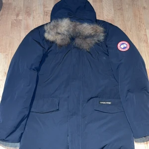 Canada Goose Jacka - Säljer en mörkblå Canada Goose Expedition Parka med huva och äkta pälsdetalj. Jackan har klassisk patch på ärmen, stora fickor framtill och robust dragkedja. Perfekt för kalla vinterdagar och riktigt ikonisk streetstil.