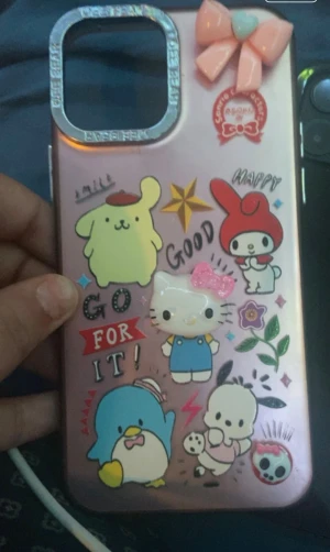 iPhone12 Skal med Sanrio Karaktärer - Sött iPhone-skal med Sanrio-figurer som Hello Kitty, My Melody och Pompompurin. Rosa design med dekorativa stickers och 3D-rosett. Skalet är i gott skick utan synliga sprickor eller större slitage, perfekt för dig som vill skydda din iPhone med stil.