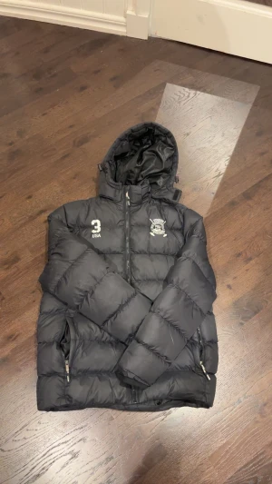 Svart pufferjacka från Vinson Polo Club - Svart pufferjacka från Vinson Polo Club med huva och dragkedja framtill. Jackan har broderad logga och siffran 3 på bröstet, samt två sidofickor med dragkedja. Perfekt för kalla dagar och har en schysst, sportig look.