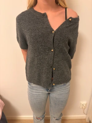Mörkgrå kortärmad stickad kofta från H&M - Mysig mörkgrå kortärmad, stickad kofta från H&M med snygga guldfärgade knappar framtill. Koftan har en loose passform och ribbade muddar vid ärmslut och nederkant. Perfekt att slänga över en t-shirt eller linne. Modellen är 158 cm och bär storlek 170. Aldrig använd förutom för foto