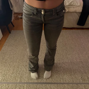 Grå bootcut jeans med låg midja - Säljer ett par gråa jeans med bootcut passform och låg midja. Jeansen har klassiska fem fickor, bälteshällor och dubbla knappar fram. Materialet är jeans och färgen är en mörkgrå ton som passar till det mesta.