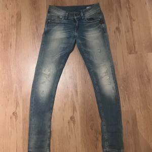 Blå slim jeans från G-Star RAW sjukt tvätt 32/32 - Snygga blå jeans från G-Star RAW med slim passform. Jeansen har slitna detaljer, klassisk femficksdesign och logotyp på fickan. Materialet är stretchigt denim för en bekväm känsla. Perfekt för dig som gillar en smal siluett och streetstil. Midja 37cm längd 109cm (78) 