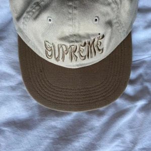 Supreme beige keps med broderi - Supreme keps i beige med brunt brätte och broderad Supreme-logga framtill i matchande ton. Justerbar snapback baktill för perfekt passform. Tillverkad i bomull och har klassisk streetwear-vibe. Perfekt accessoar för att lyfta din outfit.