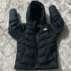 Svart pufferjacka från Nike med huva - Svart pufferjacka från Nike med klassisk logga på bröstet. Jackan har huva, dragkedja framtill och quiltad design. Perfekt för kalla dagar och har en sportig vibe. Tillverkad i syntetmaterial och har långärmad passform.