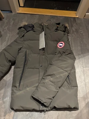 Canada goose  - Säljer en grön dunjacka från Canada Goose med klassisk patch på ärmen. Jackan har huva, dragkedja och knappar framtill samt två sidofickor med dragkedja. Perfekt för kalla dagar och riktigt snygg streetstil.