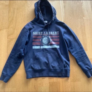Svart hoodie från Saint Laurent - Svart hoodie från Saint Laurent med tryck i rött och vitt framtill. Tröjan har huva med snörning och en stor magficka. Perfekt för dig som vill ha en avslappnad stil.