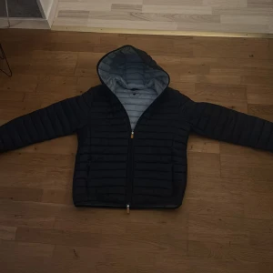 Svart pufferjacka med huva - Säljer en blå pufferjacka med huva och snygga horisontella quiltade sömmar. Jackan har dragkedja med orange detaljer och en ljusblå insida i huvan. Perfekt för kyliga dagar och har en clean, modern look. Strl L men sitter om s/m