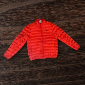 Röd dunjacka från Adidas - Säljer en röd dunjacka från Adidas med klassisk logga på bröstet. Jackan har quiltad design, lång ärm och dragkedja framtill. Materialet är lätt och smidigt, perfekt för kyliga dagar. Insidan är mörkgrå och jackan har en clean, sportig look.