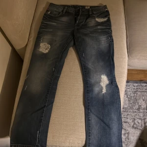 Jack and Jones  - Säljer ett par klassiska Jack and Jones jeans i mörkblå tvätt med coola slitningar och trasiga detaljer på benen. Jeansen har rak passform, fem fickor och tydliga slitningar både fram och bak för en avslappnad streetstil. Perfekt för dig som gillar vintagevibe.