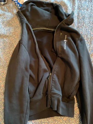 Svart Sail Racing hoodiejacka - Svart hoodiejacka från Sail Racing med dragkedja framtill och på bröstfickan. Jackan har huva och är fodrad med mjuk fleece på insidan. Perfekt för dig som gillar sportig och avslappnad stil.