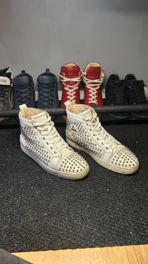 Christian Louboutin vita sneakers med nitar - Säljer ett par vita high-top sneakers från Christian Louboutin med ikonisk röd sula och silverfärgade nitar över hela skon. Skorna har snörning och är tillverkade i skinn, med rund tå och platt sula. Märkeslogga på sidan. Bra skick och skönt ingådda så väldigt bekväma. Har äkthetsbevis och kvitto som jag skickar vid intresse. Har låda, dustbags och massa andra små grejer som kom med som fås med vid köp.