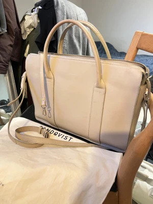 Beige Alice Sandqvist axelväska  - Snygg beige axelväska från Sandqvist i slätt skinn med stilren design. Väskan har både handtag och justerbar axelrem, samt flera fack och dragkedja upptill. Perfekt storlek för laptop och vardagsprylar, med minimalistisk look och diskreta detaljer. Små tecken på använingen om man kollar mycket noggrant. Nypris 3200. 5 Liter
