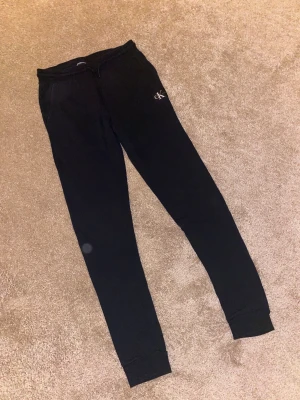 Svarta mjukisbyxor Calvin Klein Jeans - Svarta mjukisbyxor från Calvin Klein Jeans med vit CK-logga på fickan bak och mindre logga framtill. Byxorna har smal passform, ribbade muddar vid benslut och elastisk midja med snörning. Perfekta för chill eller träning.