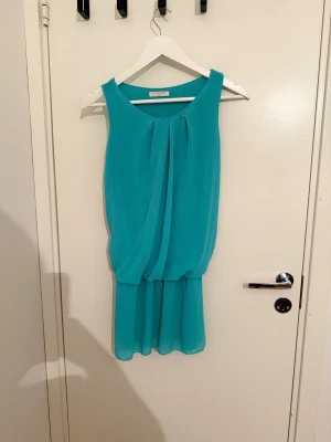 Turkos ärmlös klänning från Bobomee - Säljer en turkos ärmlös klänning från Bobomee. Klänningen har en lös passform med draperad överdel och lätt rynkad kjol. Den är kort och har rund halsringning, perfekt för dig som gillar en enkel men snygg look.