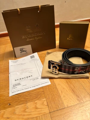 Rutig Burberry bälte med silverdetalj - Säljer ett snyggt Burberry bälte i svart och grått med röd rutig design och ikoniskt Burberry-motiv. Bältet har ett stort silvrigt 'TB'-spänne och är gjort i skinn. Perfekt accessoar för att lyfta din outfit.