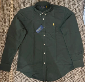 Mörkgrön skjorta från Polo Ralph Lauren - Snygg mörkgrön skjorta från Polo Ralph Lauren i klassisk passform. Skjortan har button-down krage, vita knappar och den ikoniska gula broderade loggan på bröstet. Tillverkad i mjuk bomull, perfekt för en clean och stilren look.