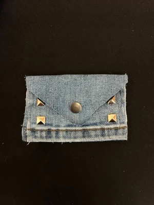 Ljusblå denim plånbok med nitar - Plånbok i ljusblå denim med fyrkantiga silverfärgade nitar på framsidan och en tryckknapp för stängning. Råa kanter ger en cool och avslappnad vibe. Perfekt storlek för kort och cash.