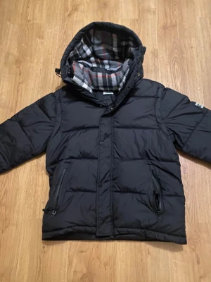 Svart pufferjacka från Burberry - Svart pufferjacka från Burberry London England med huva och rutig insida i grått, vitt och rött. Jackan har dragkedja och knappar framtill samt två fickor med dragkedja. Klassisk Burberry-patch på ärmen och quiltad design för extra värme.