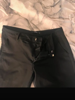 Svarta kostymbyxor med fickor - Snygga svarta kostymbyxor med klassisk passform och diskreta fickor både fram och bak. Byxorna har hällor för bälte och stängs med dragkedja och knapp. Perfekta för en stilren look och passar till skjorta eller t-shirt. LÅGMIDJADE 