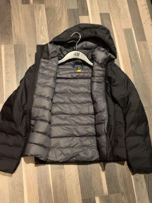 Svart pufferjacka från Polo Ralph Lauren - Snygg svart dunjacka från Polo Ralph Lauren med huva och dragkedja framtill. Jackan har quiltad insida i grått och en röd Polo-logga på bröstet. Perfekt för kalla dagar och har en clean, sportig look som funkar till streetwear.