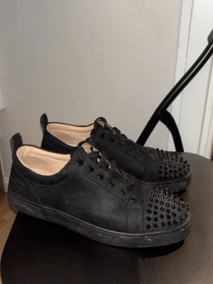Svarta loubs med nitar och röd sula - Säljer ett par svarta sneakers med coola svarta nitar på tån och snörning. Skorna har en platt sula med röd undersida och en vit innersula med mönster. Utsidan är i mocka och insidan är ljusbeige. Perfekt för dig som gillar edgy stil.