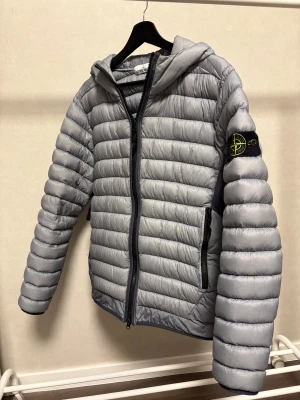 Grå dunjacka Stone Island med huva - Snygg grå dunjacka från Stone Island med huva och svarta detaljer. Jackan har quiltad design, dragkedja framtill och två sidofickor med zip. Klassisk Stone Island patch på vänster ärm. Perfekt för kyliga dagar och stilren streetwear.
