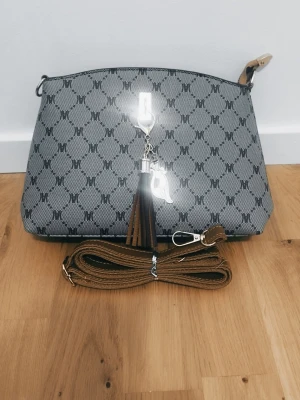 Grå axelväska med mönster och tofs - Snygg grå axelväska  med svart logomönster över hela väskan. Väskan har en avtagbar brun axelrem och en dekorativ tofs framtill i brunt. Perfekt storlek för det viktigaste och stängs med dragkedja.