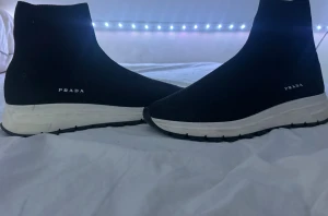 Svarta sockboots från Prada strl 40 - Säljer ett par svarta sockboots från Prada med vit tjock sula och diskret logga på sidan. Skorna är i stretchigt stickat material som sitter tight runt foten och har en rund tå. Perfekt för dig som gillar stilrena och moderna sneakers med boots-vibe. Vid snabb affär kan även sänkas ner i pris. Låda följer med