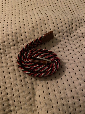Randig flätad bälte i rött och svart - Snyggt flätat bälte med randigt mönster i rött, svart och vitt. Perfekt för alla typer av byxor.  100 cm
