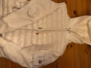 Vit Moncler Cardigan  - Säljer en snygg vit cardigan från Moncler med huva och dragkedja. Cardigan har quiltad framsida, två fickor med dragkedja och en Moncler-logga på ärmen. NFC tagen funkar. Inget fel på den. Oanvänd med. 