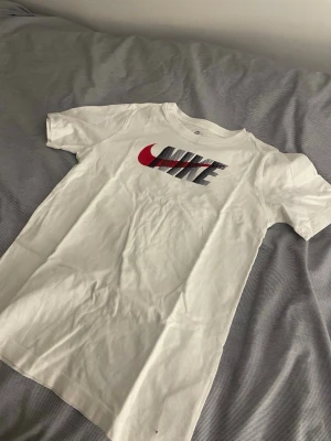 Vit Nike t-shirt med röd logga - Snygg vit t-shirt från Nike med klassisk logga i rött och svart framtill. T-shirten är i mjuk bomull och har korta ärmar och rund hals. Perfekt för en sportig och avslappnad stil.