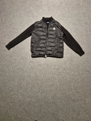 Svart pufferjacka från Moncler - Svart pufferjacka från Moncler med stickade ärmar och quiltad kropp. Jackan har dragkedja framtill, ribbad krage och broderad Moncler-logga på bröstet. Snyggt broderad detalj på ärmen. Perfekt för dig som gillar exklusiva streetwear-vibes.