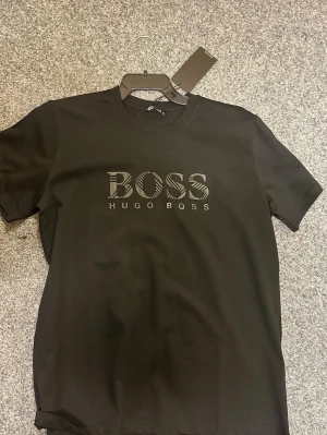 Svart BOSS t-shirt från Hugo Boss - Svart t-shirt från Hugo Boss med stort BOSS-tryck på bröstet i svart med diskreta ränder. Klassisk rund halsringning och korta ärmar. Tillverkad i mjuk bomull för en skön känsla. Perfekt för dig som gillar stilrena och snygga basplagg.