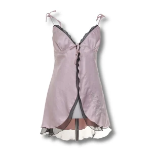 Rosa Linne - Supersöt ljusrosa linnetopp från H&M i glansig satin med svarta volangdetaljer och omlott framtill. Tunna axelband med knyt och lätt utsvängd nederkant. Perfekt för dig som gillar romantisk och feminin stil. OBS liten tråddragning (se bild)