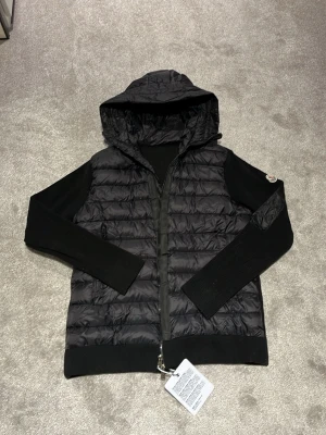 Svart Moncler cardigan med huva - Svart cardigan från Moncler med huva och ribbstickade ärmar. Jackan har quiltad framsida i syntetmaterial och dragkedja framtill. Moncler-logga på vänster ärm. Snygg, modern look med blandning av material och detaljer.