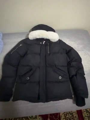 Svart pufferjacka Moose Knuckles - Svart pufferjacka från Moose Knuckles med vit fuskpäls på huvan och ikoniska metallogo på ärmen. Jackan har dragkedja, flera fickor och en quiltad design som håller dig varm. Perfekt för kalla vinterdagar och har en riktigt clean streetstil.