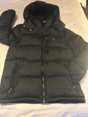 Svart dunjacka från Ralph Lauren - Svart dunjacka från Ralph Lauren med huva och dragkedjor framtill. Jackan har en röd broderad logga på bröstet och flera praktiska fickor med dragkedja. Perfekt för kalla dagar och riktigt snygg till streetwear-stilen. Storlek XL i barn [xs-S i herr] skriv vid frågor!