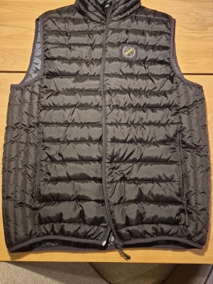 Svart dunväst från Norra Stå - Svart quiltad dunväst från Norra Stå med gul broderad logga på axeln och klubbmärke på bröstet. Västen har dragkedja framtill och två sidofickor med dragkedja. Perfekt för lager-på-lager och har en clean, sportig look. Storlek M