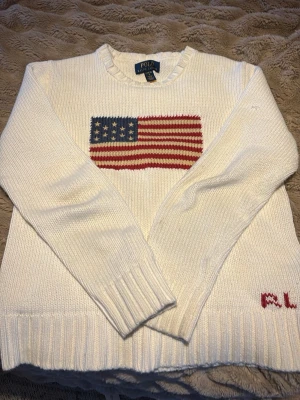 Vit stickad tröja Polo Ralph Lauren - Vit stickad tröja från Polo Ralph Lauren med amerikansk flagga broderad på bröstet och RL-broderi i rött vid nederkanten. Rund hals, ribbade muddar och långärmad. Perfekt för dig som gillar klassisk och clean stil.
