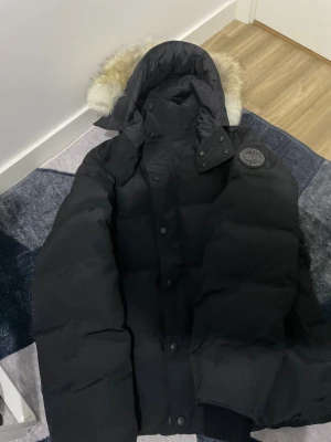 Svart dunjacka från Canada Goose - Säljer en svart dunjacka från Canada Goose med stor huva och fluffig pälsdetalj. Jackan har patch med logga på ärmen, dragkedja och knappar framtill. Perfekt för kalla vinterdagar och riktigt snygg streetstil.