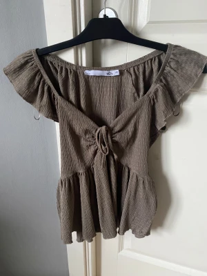 Brun volangblus med knyt XS - Supersöt brun blus från 157 i storlek XS. Toppen har volangärmar, smockad struktur och ett snyggt knyt framtill. Offshoulder-modell som ger en chill vibe och flowig passform. Perfekt till jeans eller kjol för en trendig look.