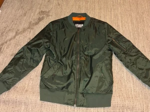 Grön bomberjacka Urban Classics XL - Snygg mörkgrön bomberjacka från Urban Classics i storlek XL. Jackan har orange foder, ribbade muddar och dragkedja framtill. Klassisk bomberstil med fickor och en extra ficka med dragkedja på ärmen. Perfekt för dig som gillar streetwear. Strlk XL men passar L bättre.