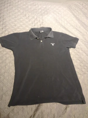 Svart piké från GANT med krage - Svart pikétröja från GANT med klassisk krage och två knappar framtill. Diskret GANT-logga broderad på vänster bröst. Modellen är kortärmad och har en slim passform som sitter snyggt på kroppen. Materialet är mjuk bomull som andas bra.