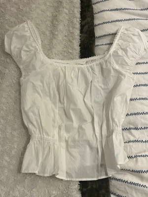 Vit blus med volang och puffärm - Säljer en söt vit blus med puffiga korta ärmar och volang nedtill. Blusen har en rundad hals med spetsdetalj och är lätt rynkad för en romantisk vibe. Perfekt till jeans eller kjol för en fräsch look. 