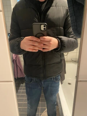 Svart pufferjacka från H&M - Svart pufferjacka från H&M med klassisk krage och dragkedja framtill. Jackan har quiltad design, två sidofickor och långa ärmar med elastiska muddar. Perfekt för kalla dagar och enkel att matcha med jeans eller hoodies.