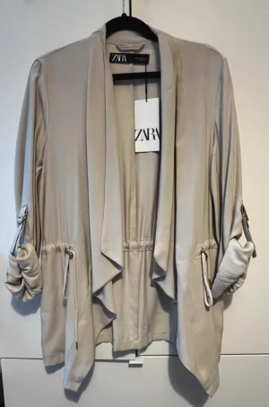 Beige cardigan jacka från Zara - Snygg beige cardigan jacka från Zara med avslappnad passform och dragsko i midjan. Jackan har långa ärmar som kan rullas upp och fästas med knapp, samt öppen framtill för en chill look. Tillverkad i mjukt material med viskos och polyester.