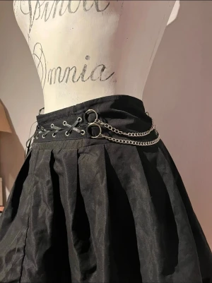 Svart kortkjol med kedjor och snörning - Säljer en svart kortkjol med volangkant, snörning och silverfärgade kedjor i midjan. Kjolen har veckad design och dragkedja i sidan.
