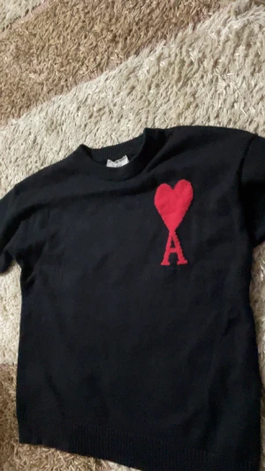 Svart stickad tröja från Ami Paris - Svart stickad tröja från Ami Paris med röd hjärtapplikation och bokstaven A på bröstet. Tröjan har rund hals och långa ärmar, tillverkad i mjukt material som passar perfekt för kyliga dagar. Snygg och stilren design med ikonisk Ami-logga.
